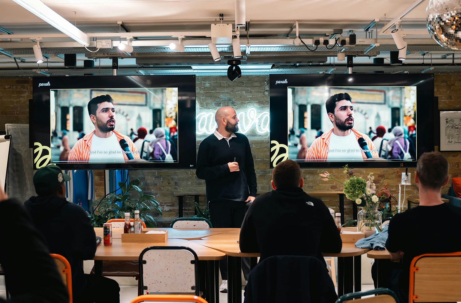 Man presenting in front of dual screens displaying video content, showcasing commercial AV technology in a modern meeting space, with audience engaged in a business setting.